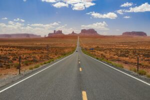 A long, empty highway stretches through Monument Valley under a bright blue sky.